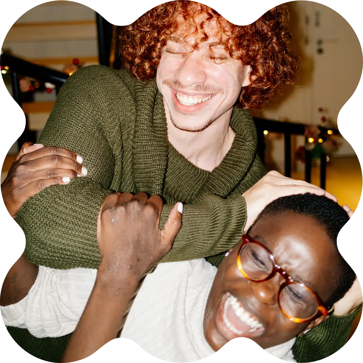 Two friends laughing and playfully embracing indoors, sharing marijuana facts—one wearing glasses, the other in a green sweater.