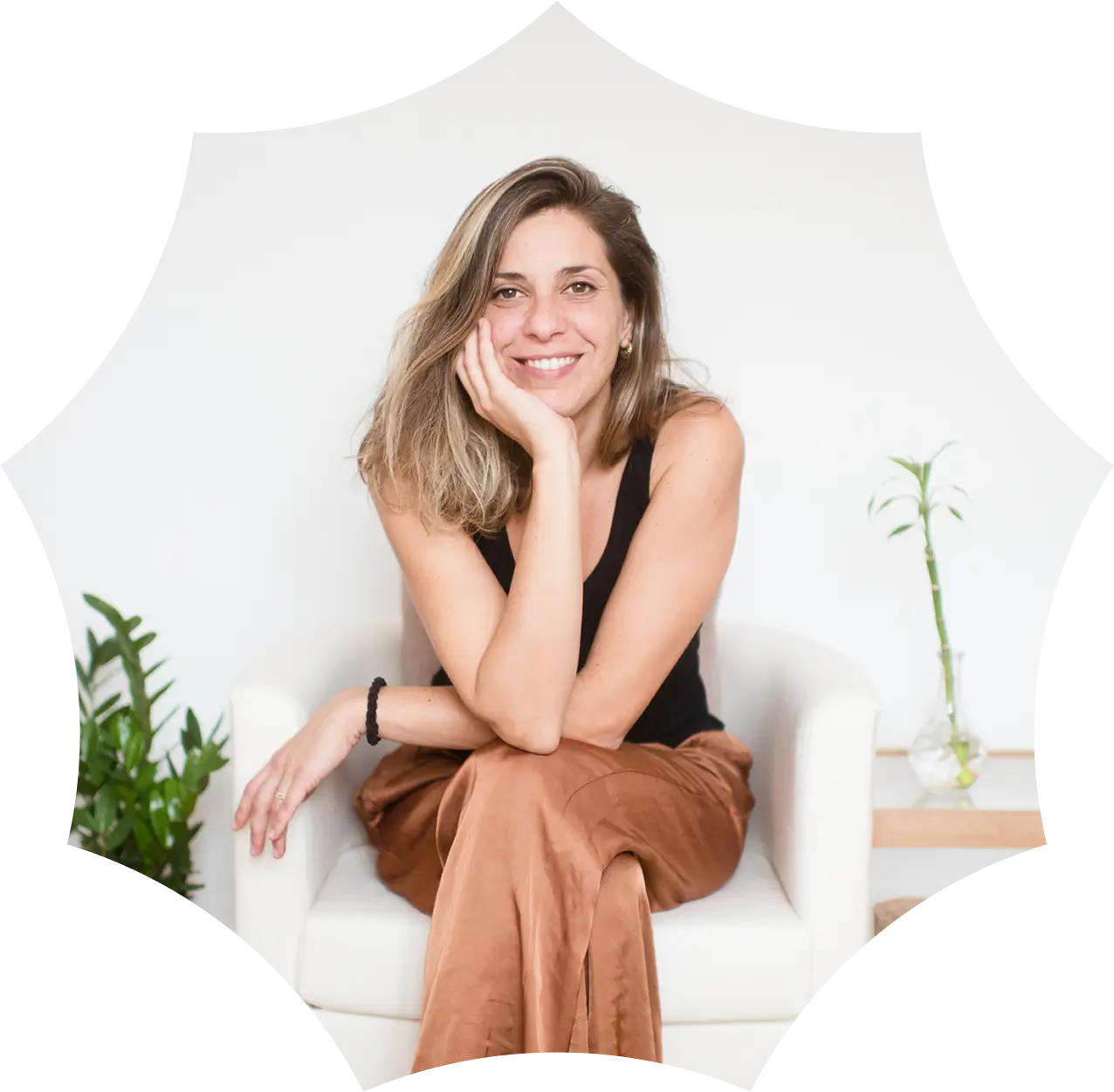 A woman with long hair, wearing a black top and brown pants, sits smiling on a white chair beside green plants—reflecting the natural side of medical marijuana in CT.