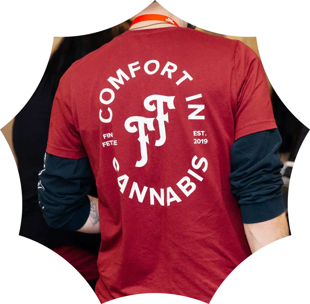 Person wearing a red shirt with white text on the back that says COMFORT IN CANNABIS, “FF,” and “EST. 2019,” at a MA medical dispensary, with others blurred in the background.