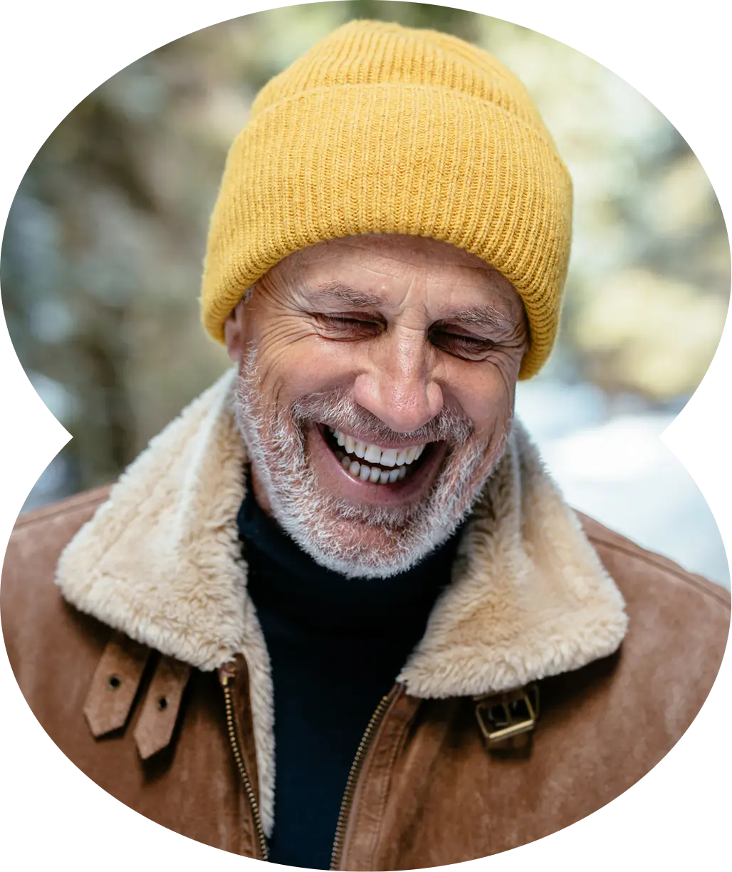 An older man with a white beard, wearing a yellow knit beanie and brown shearling jacket, smiles outdoors while discussing a small business partnership agreement.