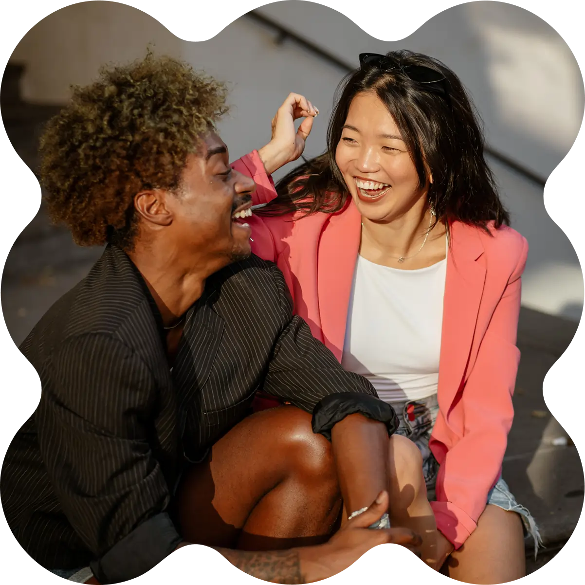 A man and woman sit on outdoor steps, smiling and laughing together as they chat about cannabis products. The photo features a scalloped black border.
