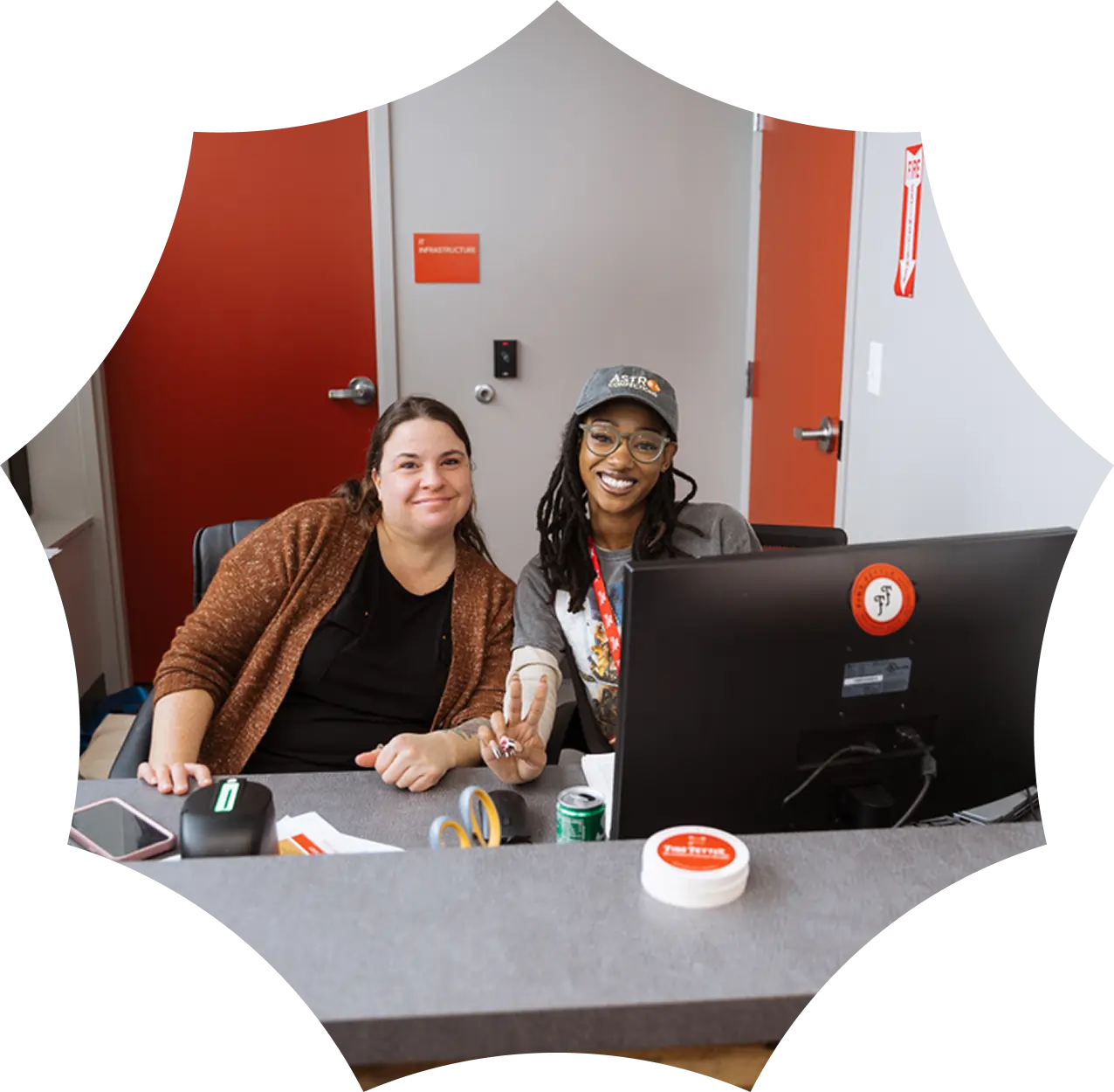 Two women smiling while sitting at a desk with a computer and office supplies in a cannabis dispensary, with a red door in the background.