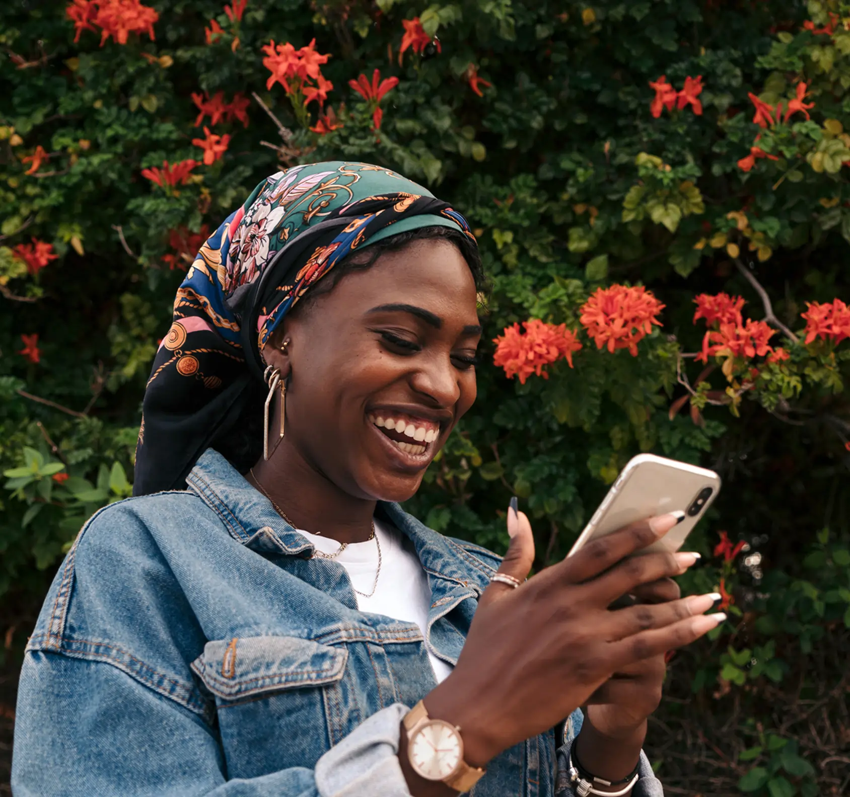A smiling woman in a headscarf and denim jacket checks her smartphone, standing outside a cannabis dispensary framed by green bushes with red flowers.