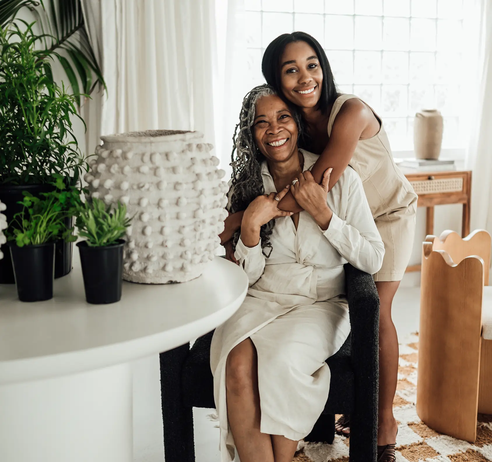 A smiling young woman stands behind and hugs an older woman seated in a bright, modern room with plants, neutral decor, and touches inspired by a Georgia dispensary.