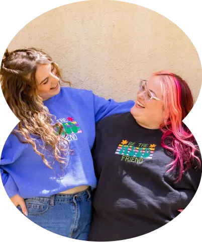 Two people smile at each other outside a cannabis dispensary, wearing sweatshirts that say Be the friend with colorful designs.