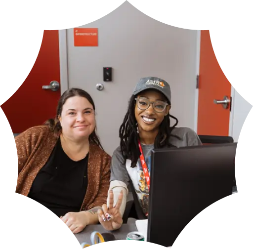 Two women sit together at a desk in a cannabis dispensary, smiling at the camera. One flashes a peace sign. A computer monitor and office doors are visible in the background.