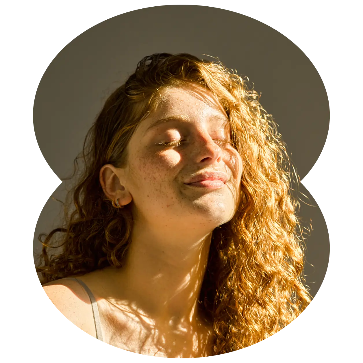 A woman with curly red hair and freckles closes her eyes and smiles softly, enjoying sunlight on her face outside a Massachusetts cannabis dispensary.