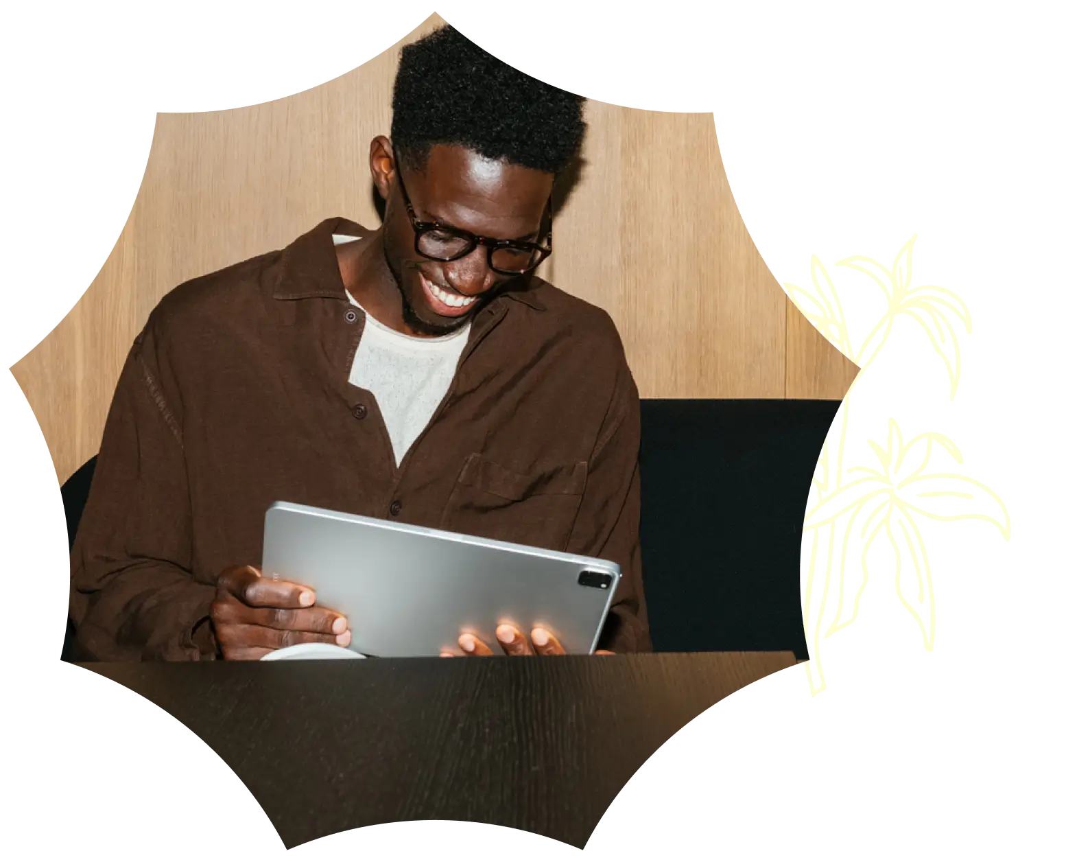 A person smiling while looking at a tablet, sitting at a dark table with a light wooden wall and a white plant illustration—exploring medical marijuana in CT.