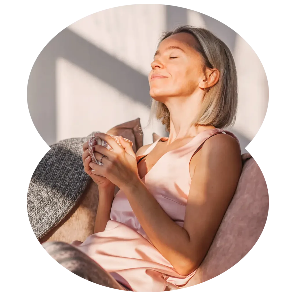 A woman with short blonde hair sits on a couch, smiling with her eyes closed, holding a mug and enjoying the sunlight as she reads about marijuana facts.