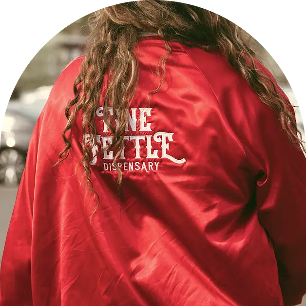 Person with long, wavy hair wearing a red jacket that says FINE FETTLE DISPENSARY on the back, standing outdoors near a cannabis dispensary.