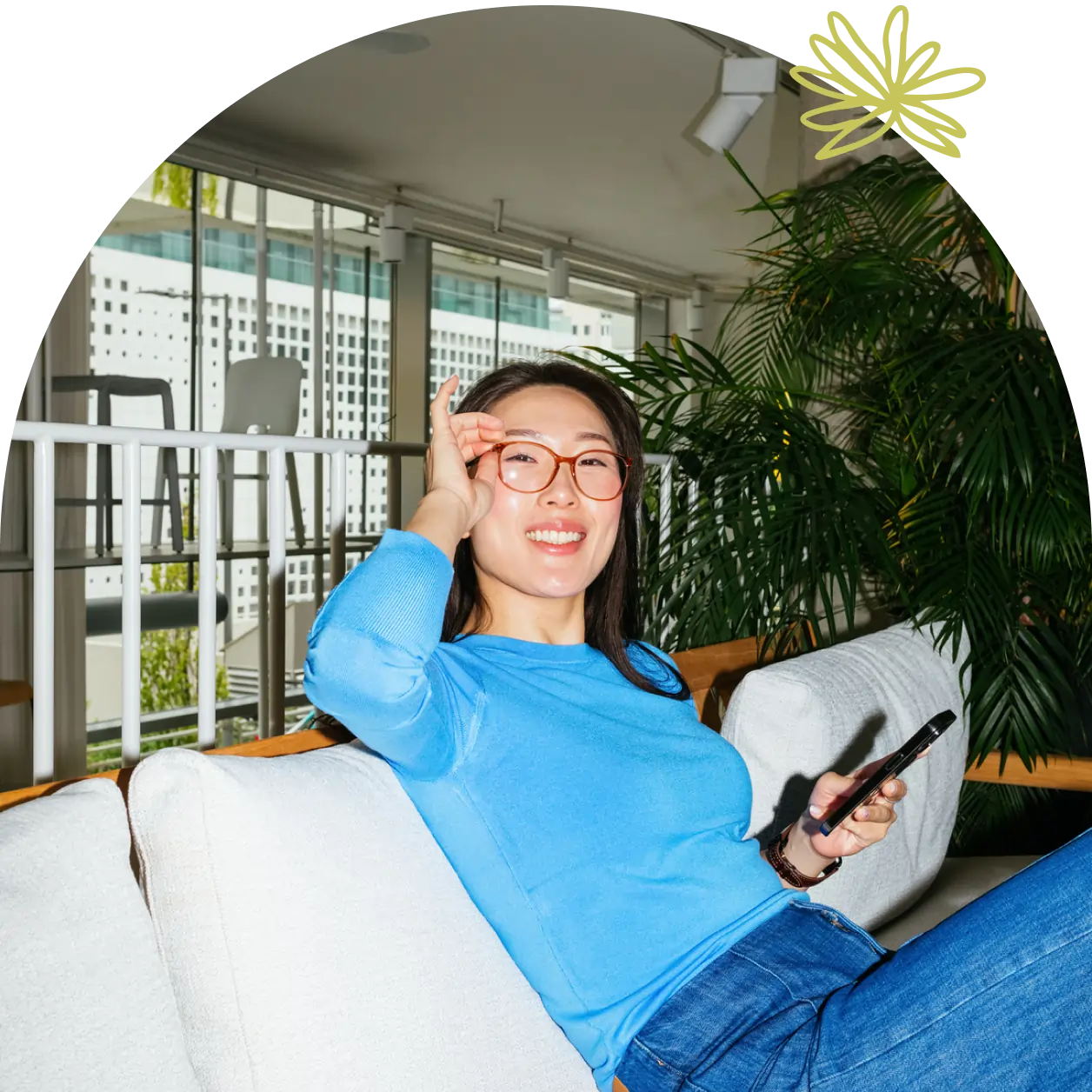 A woman in a blue sweater sits on a couch, smiling and holding a smartphone, possibly reviewing a small business partnership agreement, with greenery and large windows in the background.