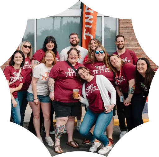 A group of ten people pose and smile outside a building, wearing FINE FETTLE shirts in maroon and white—outside their local cannabis dispensary. One person in the center holds a beverage.