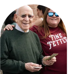 An older man holding cash and smiling stands next to a woman in sunglasses and a FINE FETTISH shirt, both looking happy outdoors by a cannabis dispensary.