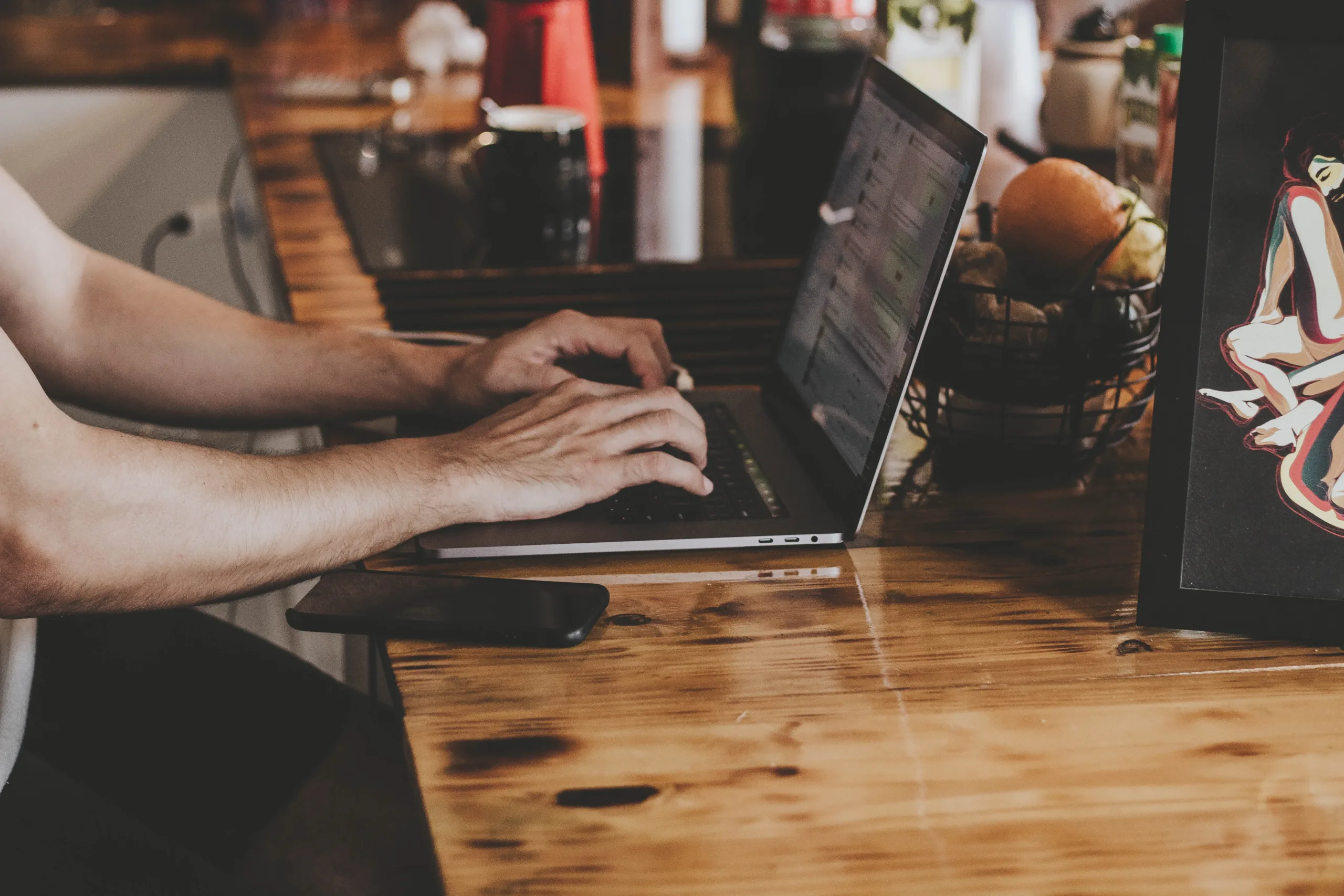 Person typing on a laptop at a wooden table with a phone nearby and a fruit basket and a framed picture in the background.