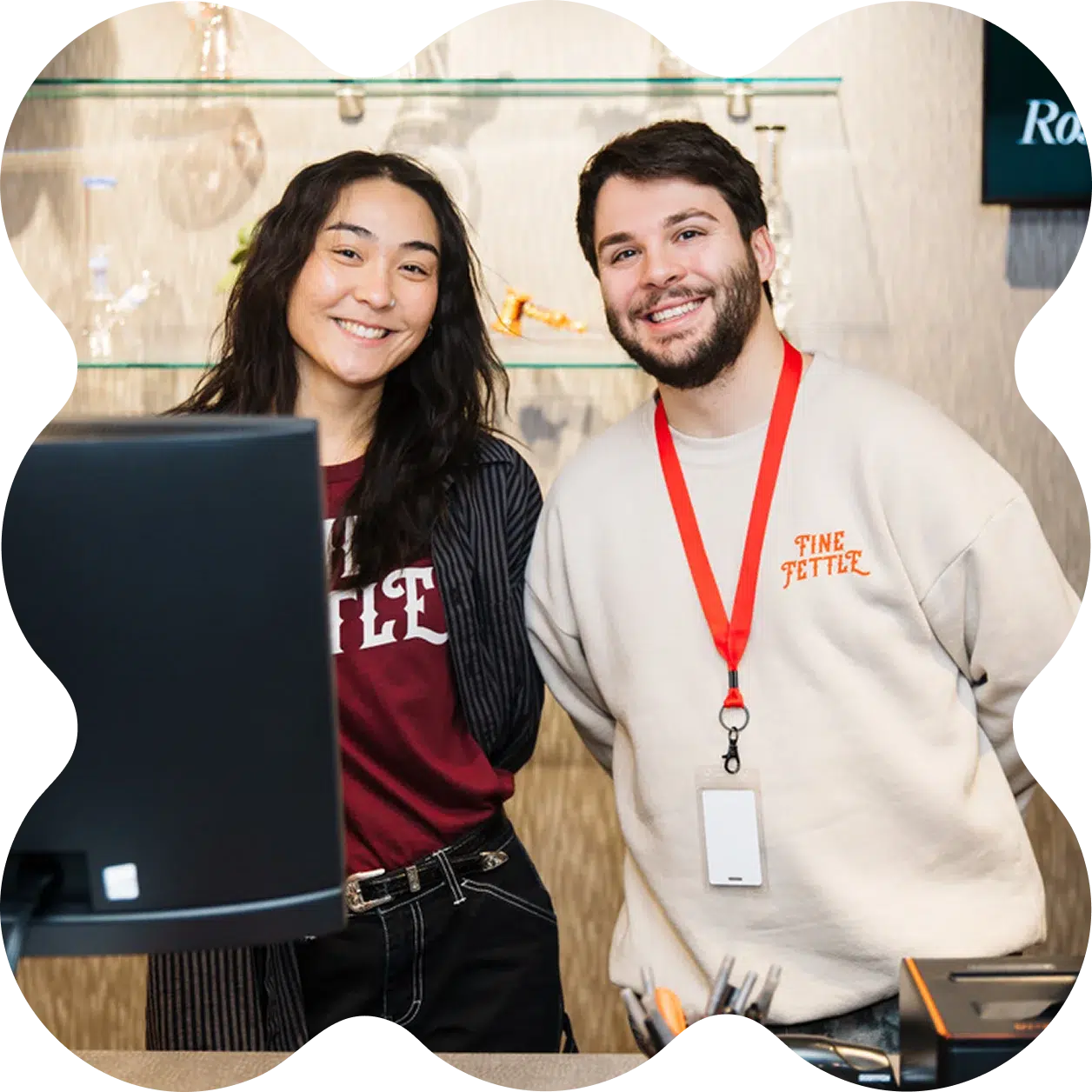 Two people smiling behind a cannabis dispensary reception desk—one in a beige sweatshirt with a red lanyard, the other in a maroon shirt and striped jacket.