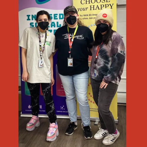 Three people wearing face masks and casual clothes stand together indoors in front of colorful banners with positive messages.