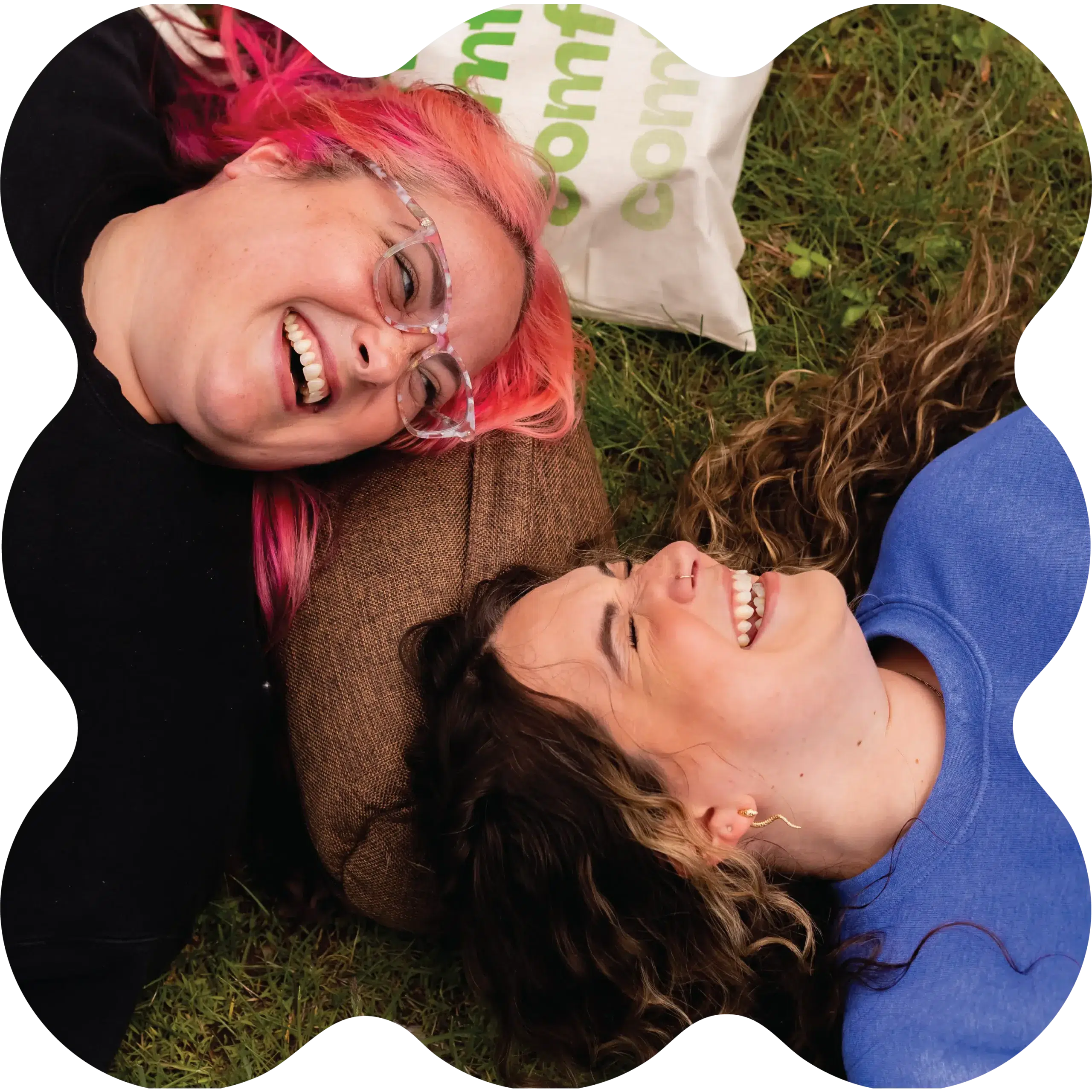 Two women lying on the grass outside a CT cannabis dispensary, heads together, smiling and laughing—one with pink hair and glasses, the other with wavy brown hair.