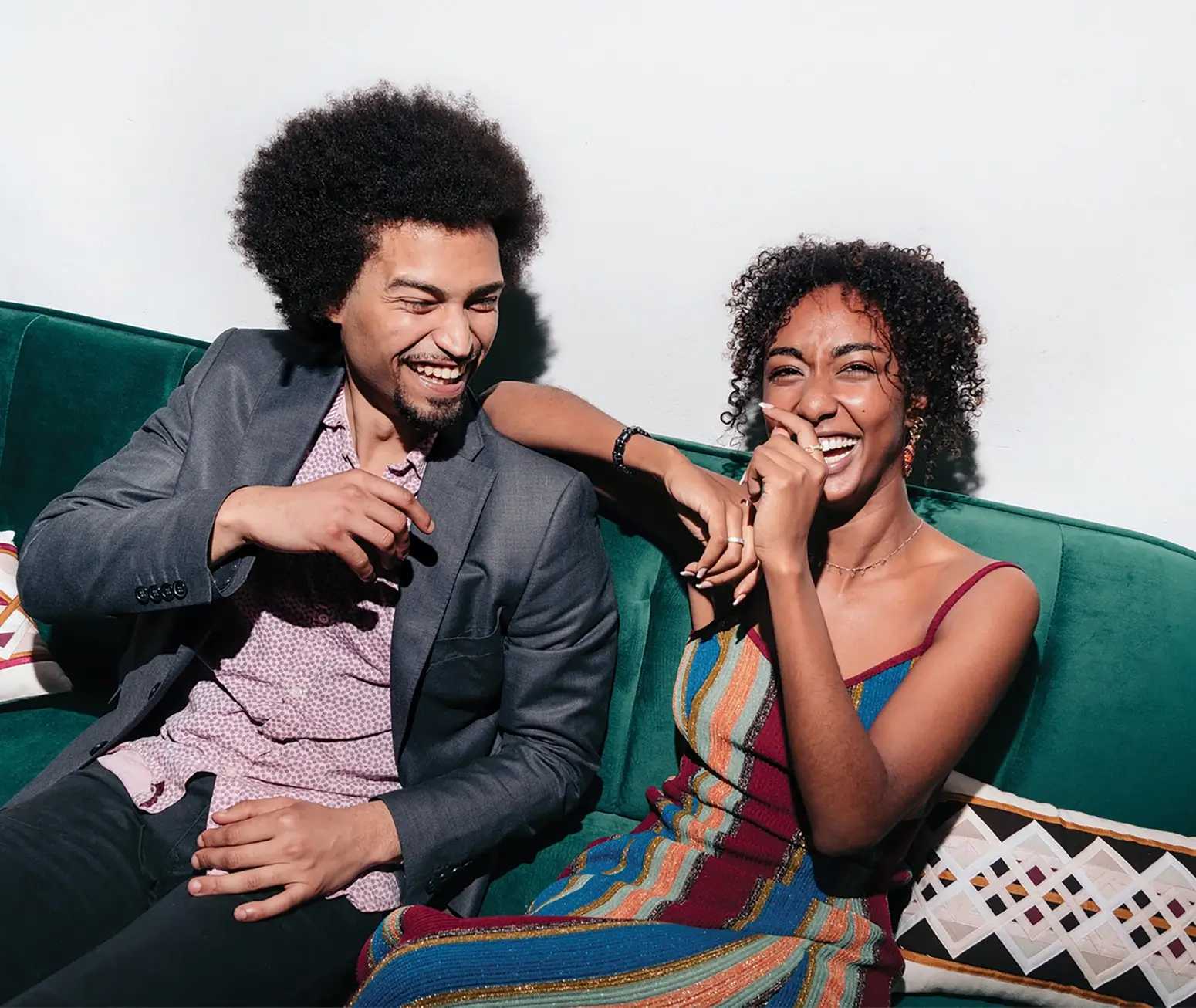 Two people sitting on a green couch, smiling and laughing together, one wearing a gray blazer and the other a colorful striped dress.