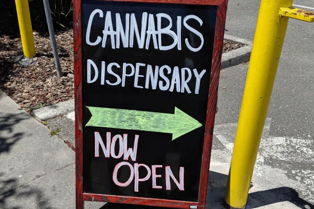 A sidewalk sign reads: CANNABIS DISPENSARY with a green arrow pointing left and NOW OPEN in red letters at the bottom.