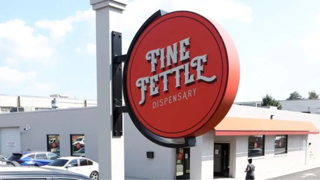 A large red sign reading FINE FETTLE DISPENSARY stands outside a modern, single-story building with parked cars and a person walking nearby.