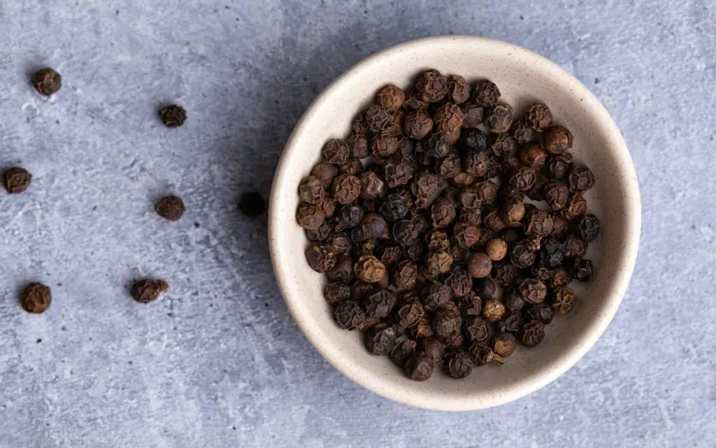 A white bowl filled with whole black peppercorns sits on a textured gray surface, with a few peppercorns scattered nearby.