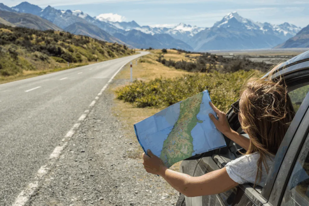 A woman hangs out the driver's window of a car holding a map and looking at an open road.