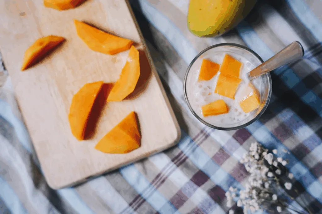 A glass of milk with mango cubes and a spoon sits beside a cutting board with sliced mango on a checkered cloth.