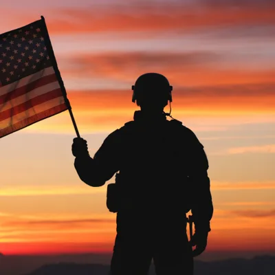 Silhouette of a soldier holding an American flag against a colorful sunset sky, honoring Veteran Cannabis Access with free Veterans Day medical cannabis evaluations.
