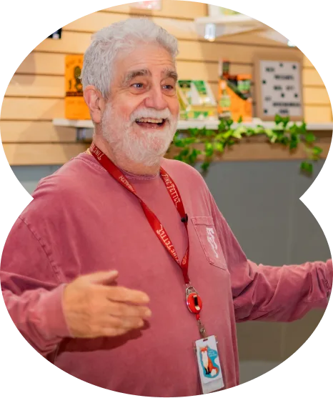 An older man with gray hair and beard smiles warmly, gesturing with open arms in a cannabis dispensary, with wooden shelves and plants in the background.