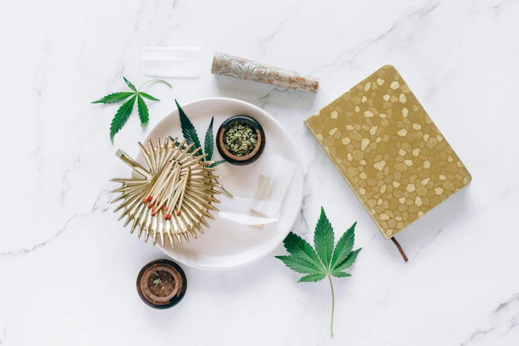 A tray with rolled joints, cannabis leaves, a grinder with cannabis, rolling papers, and a patterned notebook on a white marble surface.