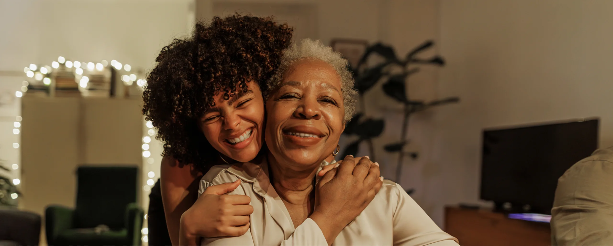A young woman hugs an older woman from behind, both smiling warmly in a cozy, softly lit living room with blurred lights in the background.