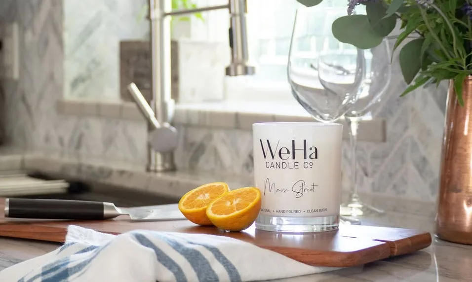 A white WeHa Candle Co. candle sits on a kitchen counter next to a sliced orange, a knife, a towel, and a vase of flowers.