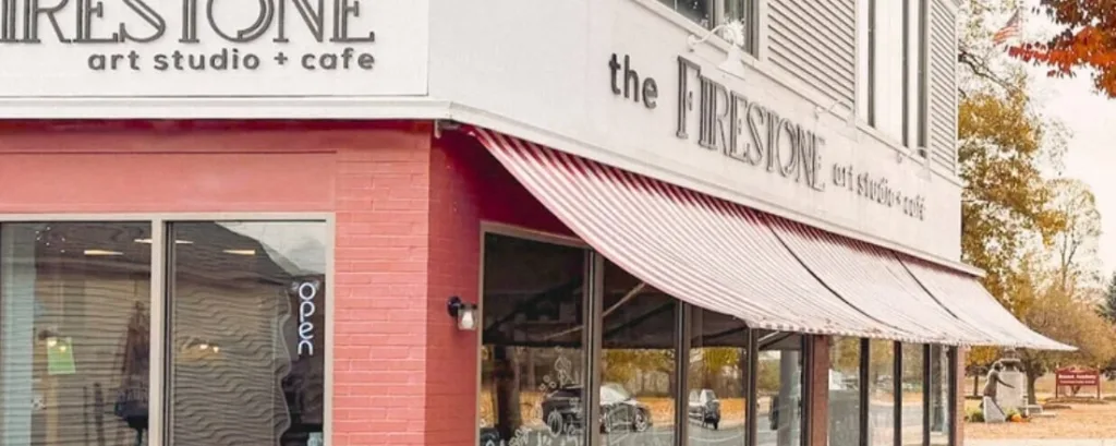 Street view of The Firestone Art Studio + Cafe with large windows, a red-and-white striped awning, and autumn trees in the background. An open sign hangs in one window.