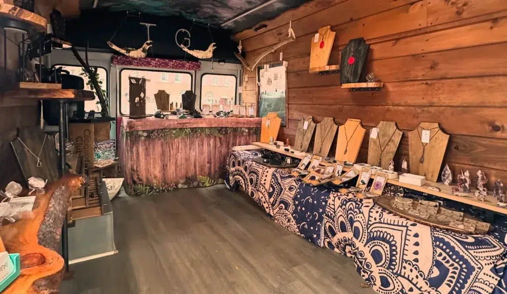 A small shop with wooden walls displays handcrafted jewelry on tables and stands, with rustic decor and natural light from a window at the back.