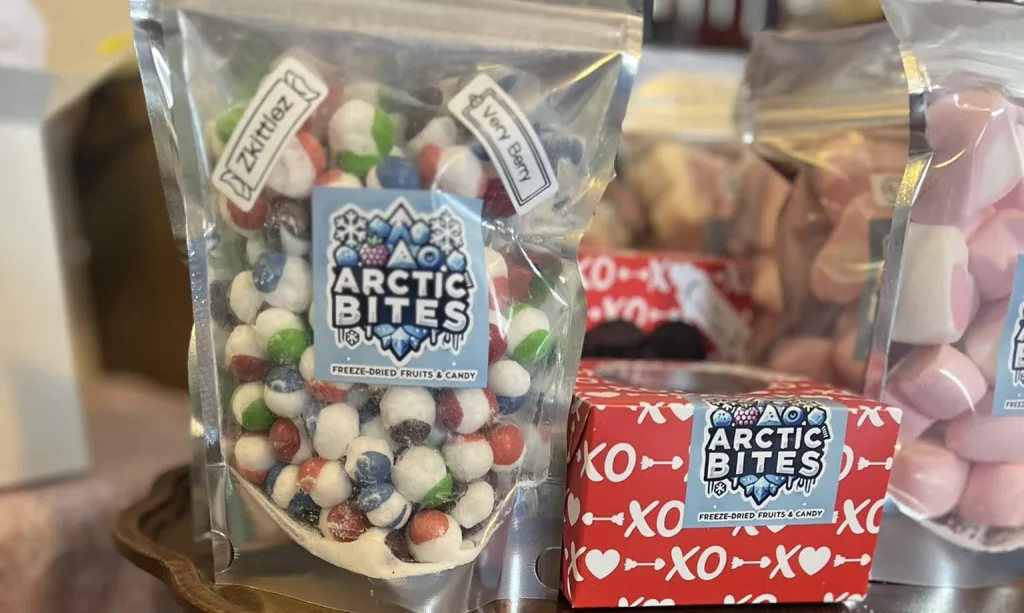 A clear bag of Arctic Bites freeze-dried fruits and candy stands next to a red box with white XOXO print and pink candies in the background.