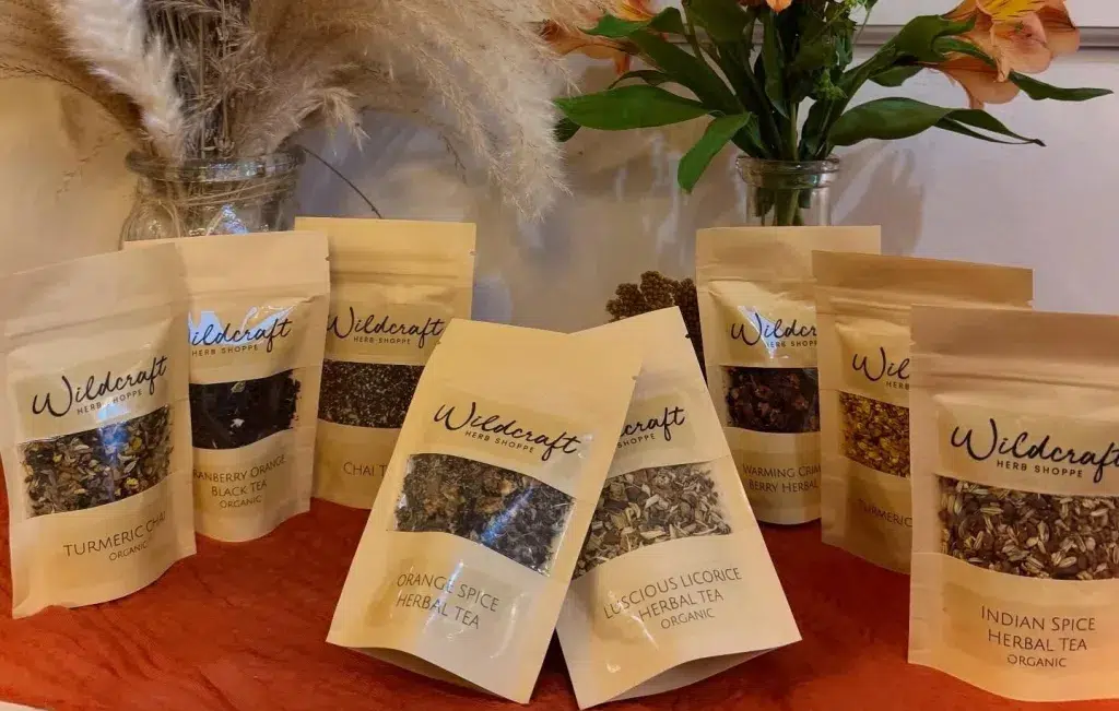 Bags of Wildcraft Herb Shop herbal teas are arranged on a table, with vases of flowers and pampas grass in the background.