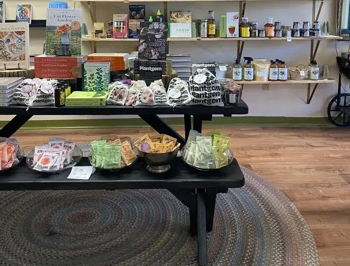 A display table with seed packets, gardening books, and small gift items set on a round rug in a shop with shelves of jars and products in the background.