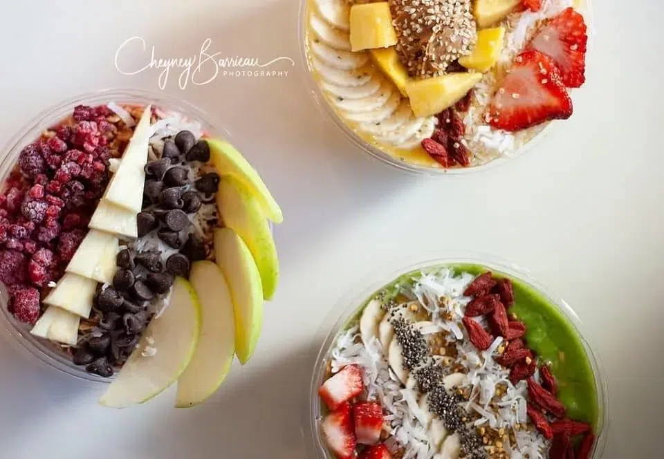 Three colorful smoothie bowls topped with a variety of fresh fruits, coconut flakes, seeds, chocolate chips, and other healthy toppings are arranged on a white surface.