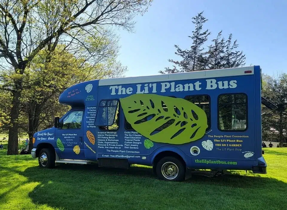 A blue bus labeled The Lil Plant Bus with green leaf graphics is parked on grass near trees under a sunny sky.