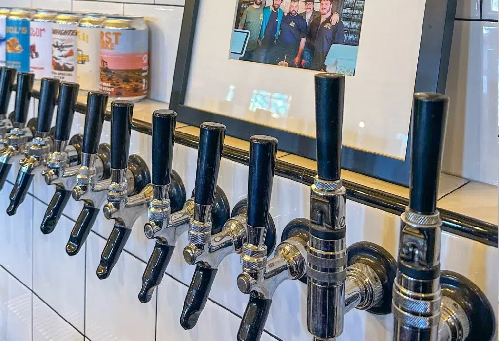 Row of beer taps mounted on a tiled wall, with a framed photo of a group of people and some beer cans displayed above the taps.