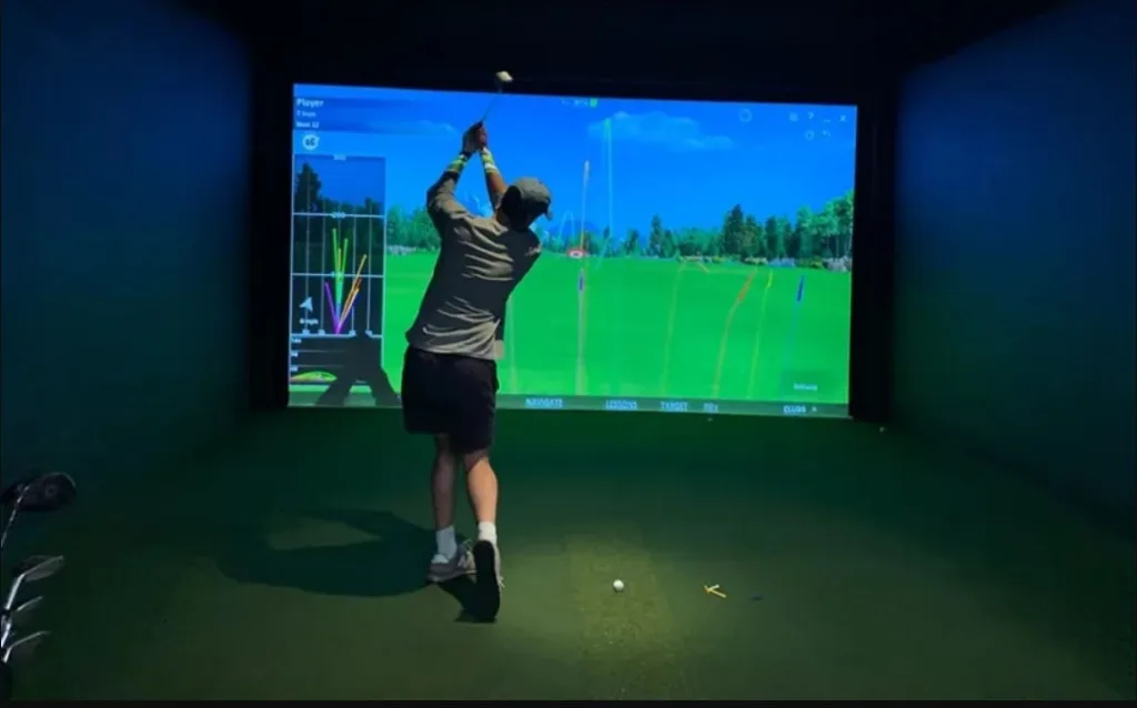 Person swinging a golf club indoors towards a large screen displaying a virtual golf course and ball trajectory data.