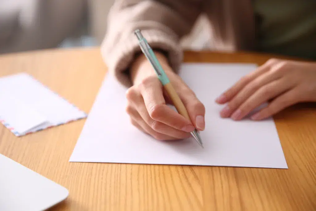 A person holding a pen and writing on a blank sheet of white paper at a wooden table.