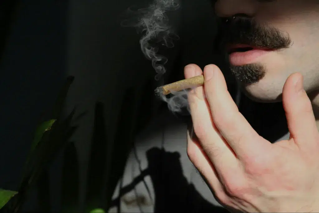 A man taking a hit off a cannabis joint as smoke emits from the tip.