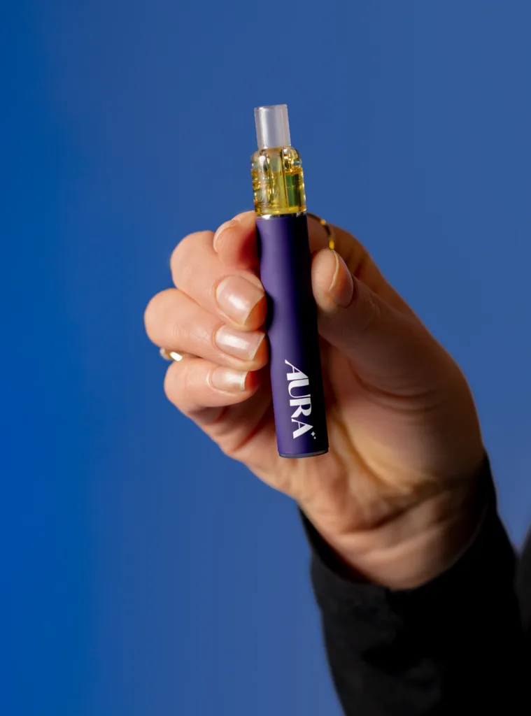 A hand holding a purple vape pen labeled AURA with a clear cartridge, against a solid blue background.