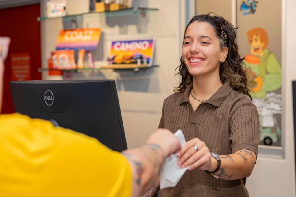 A smiling cashier hands a receipt to a customer at a counter, with colorful signs and merchandise displayed in the background.