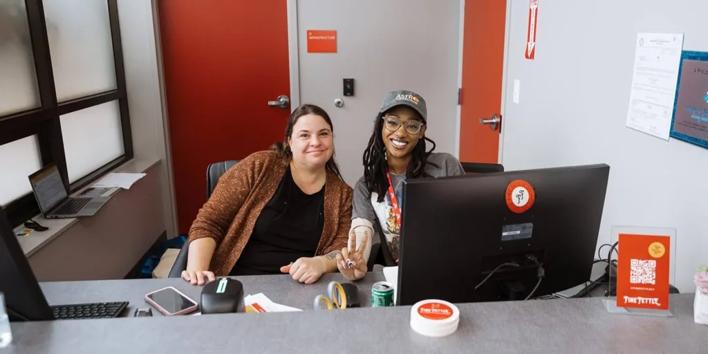Two women sitting at an office desk, smiling at the camera. They are surrounded by office supplies and a computer in a modern workspace with red doors and grey walls.