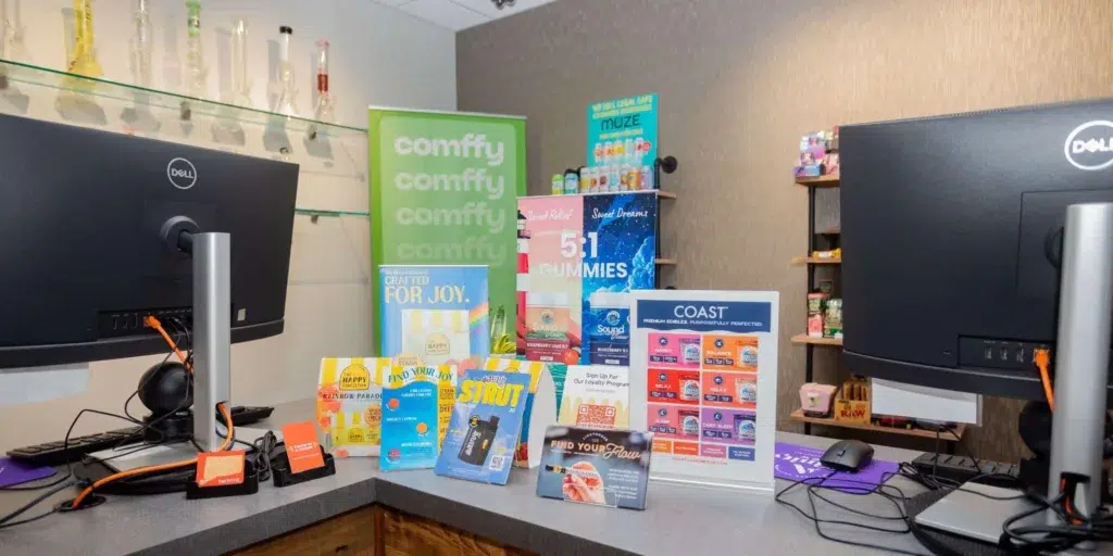 A retail counter with two monitors, colorful CBD and wellness product displays, and glass pipes on shelves in the background.
