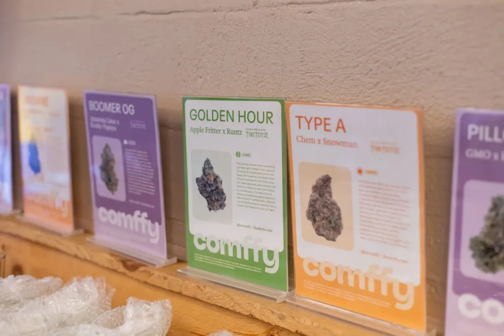 Four cannabis product displays with names, strain info, and images of buds stand on a wooden shelf against a beige wall; signs are labeled Boomer OG, Golden Hour, Type A, and more.