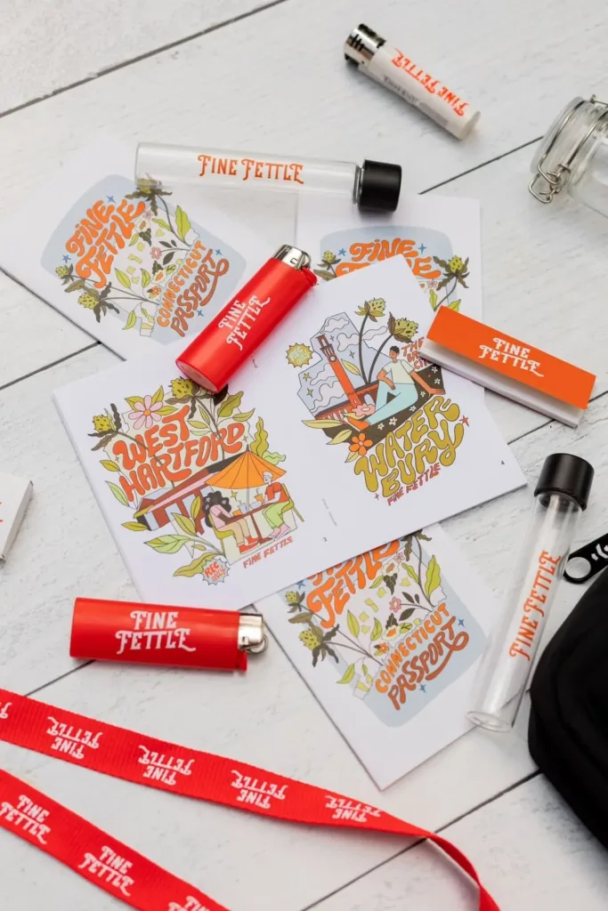 Colorful postcards and red lighters labeled Fine Fettle are arranged on a white wooden surface, along with a red lanyard, matchboxes, jars, and a black bag.