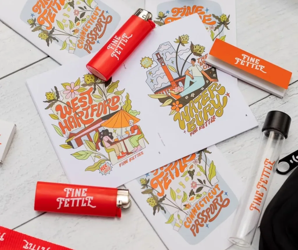 Colorful postcards and red lighters labeled Fine Fettle are arranged on a white wooden surface, along with a red lanyard, matchboxes, jars, and a black bag.